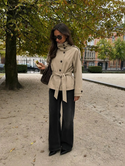 Woman wearing the Blair Belted Waist Cropped Trench Jacket in Khaki, styled with wide-leg black trousers and black heels, photographed outdoors in an autumn setting